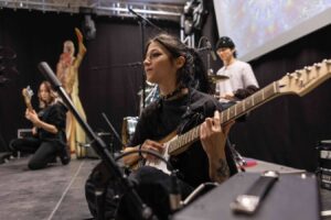 Lehrlingsportal.at Drei Musiker treten auf der Bühne auf; eine Frau spielt im Vordergrund E-Gitarre, während im Hintergrund eine andere Gitarristin und ein Schlagzeuger zu sehen sind.