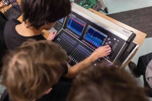 Lehrlingsportal.at Drei Personen bedienen ein digitales Audiomischpult mit mehreren Schiebereglern und Touchscreens, die von oben betrachtet werden.